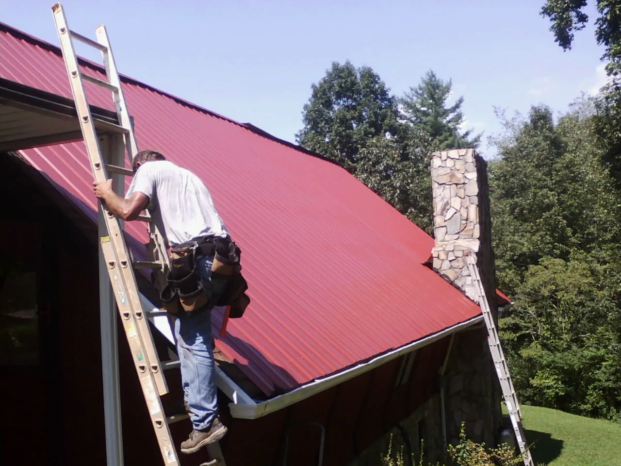 Roofer on a ladder beside a red metal roof and stone chimney — Soffit & Fascia Repair by Roofing Brothers in Loganville